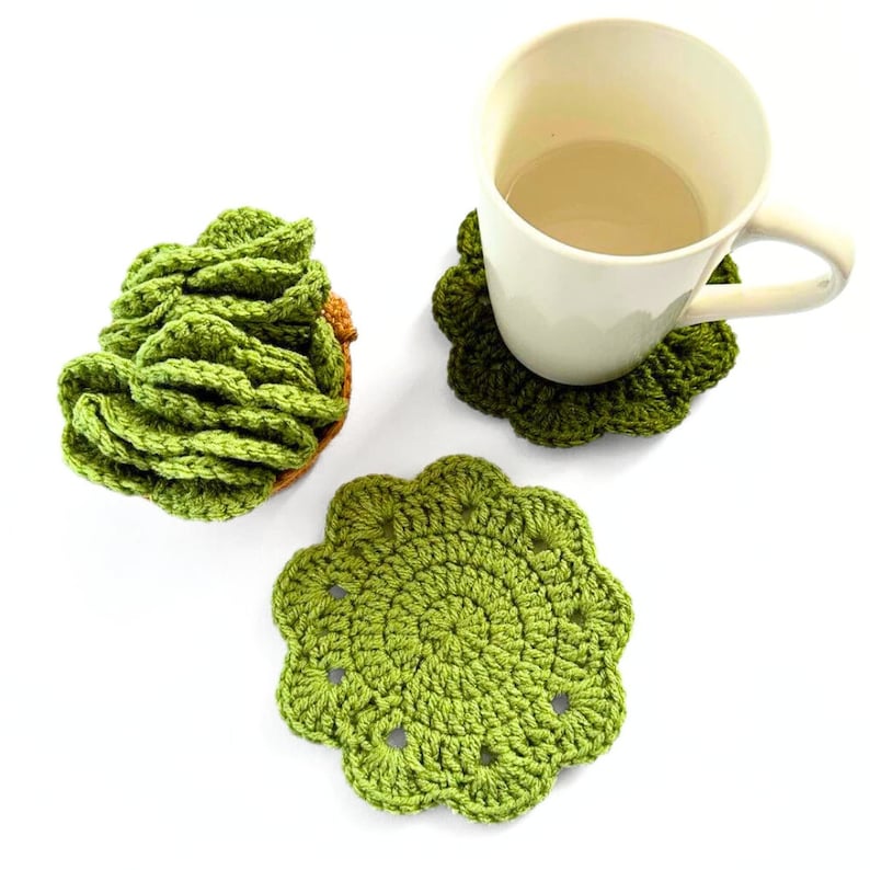 May include: Three green crocheted coasters, one shaped like a flower and two shaped like a swirl. A white mug with a light brown beverage sits on one of the coasters.