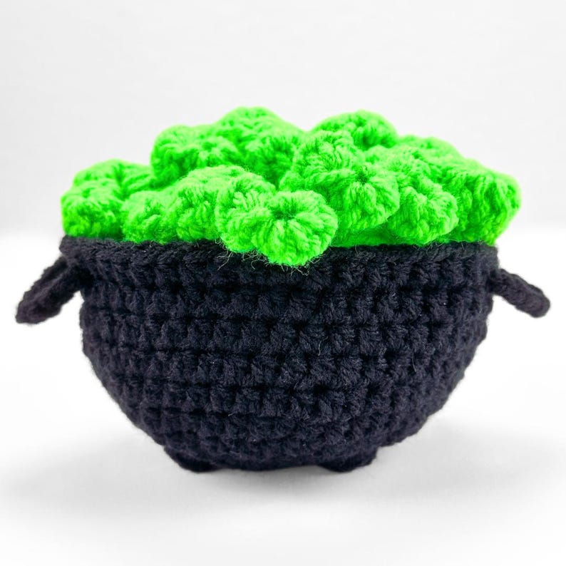 May include: A black crocheted cauldron with two small side handles, filled with a cluster of bright neon green crocheted bubbles or foam that extends above the rim of the pot, creating a textured and rounded appearance in a soft yarn material.