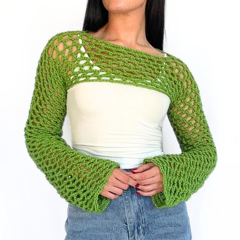 May include: A green crochet shrug with long sleeves and a wide, open neckline. The shrug is made with a delicate, open stitch pattern.