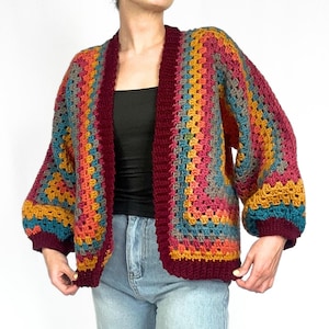 May include: A colorful crochet cardigan with a granny square pattern in shades of teal, orange, yellow, pink, and burgundy. The cardigan has a relaxed fit and long sleeves.