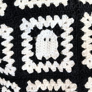 May include: A crochet textile featuring a repeating pattern of black and off-white granny squares. One central square displays a white ghost motif with two dark eye holes, integrated into the center of the square surrounded by black yarn.