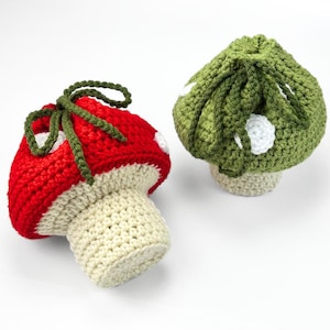 May include: Two crocheted mushroom-shaped pouches with drawstring closures. One pouch features a bright red cap with white spots and a cream-colored stem, while the other has an olive green cap with white spots and a matching olive green stem.