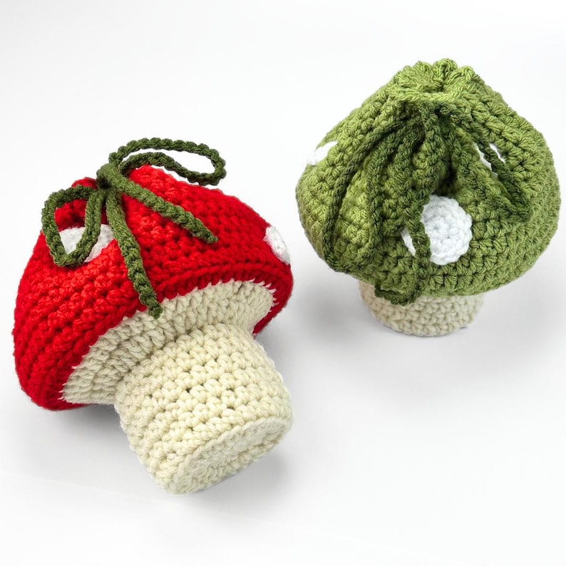 May include: Two crocheted mushroom-shaped pouches with drawstring closures. One pouch features a bright red cap with white spots and a cream-colored stem, while the other has an olive green cap with white spots and a matching olive green stem.