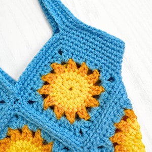 May include: A crochet top constructed from square motifs featuring a central sun design. The sun pattern uses vibrant yellow and orange yarn, while the surrounding square border is made of bright blue yarn. The piece has a thick shoulder strap.