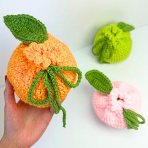 K&ouml;nnte beinhalten: Drei runde, geh&auml;kelte Beutel in Fruchtform mit Kordelzug. Ein Beutel ist orange mit einem gr&uuml;nen Blatt, einer ist limettengr&uuml;n mit einem gr&uuml;nen Blatt und einer ist hellrosa mit einem gr&uuml;nen Blatt. Jeder Beutel hat eine strukturierte Oberfl&auml;che.