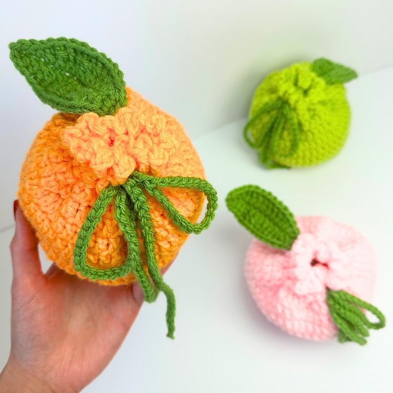 K&ouml;nnte beinhalten: Drei runde, geh&auml;kelte Beutel in Fruchtform mit Kordelzug. Ein Beutel ist orange mit einem gr&uuml;nen Blatt, einer ist limettengr&uuml;n mit einem gr&uuml;nen Blatt und einer ist hellrosa mit einem gr&uuml;nen Blatt. Jeder Beutel hat eine strukturierte Oberfl&auml;che.