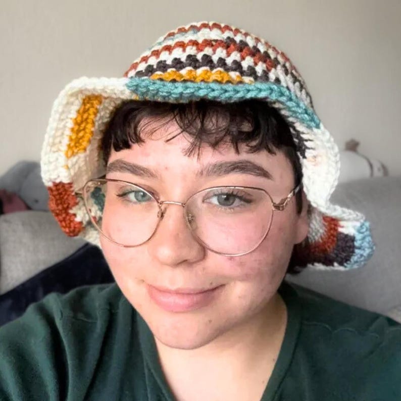 May include: A crochet bucket hat featuring a multi-coloured striped pattern in shades of cream, mustard yellow, rust orange, dark brown, and light blue. The hat has a wide, flexible brim and a textured, hand-stitched appearance throughout the design.