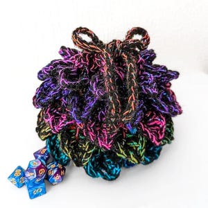 May include: A round, crocheted drawstring pouch featuring a layered, petal-like texture in black yarn with iridescent purple, blue, and green metallic highlights. Several multi-colored, translucent polyhedral dice are scattered at the base.