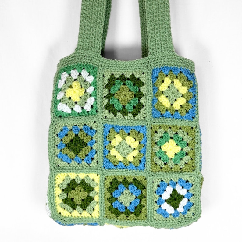 May include: A handmade crochet tote bag constructed from nine square granny squares in shades of sage green, forest green, bright yellow, sky blue, and white. The bag features two sturdy crocheted handles and a consistent green yarn border.
