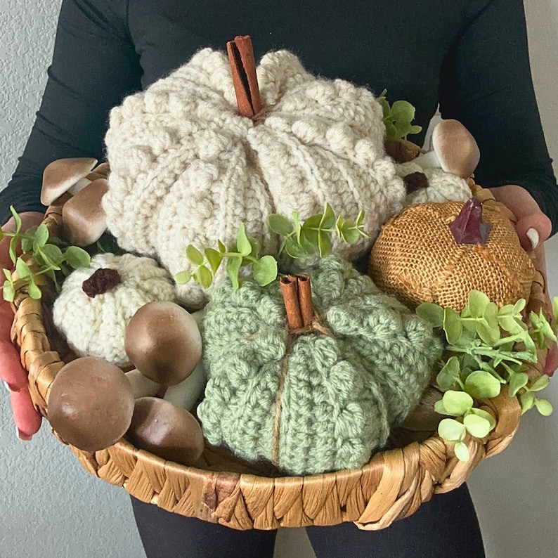 Puede incluir: Una cesta tejida que contiene varias calabazas de ganchillo en color crema y verde salvia, una calabaza dorada texturizada y figuras de setas marrones. Las calabazas tienen tallos de canela y ramas de eucalipto verde como adorno.