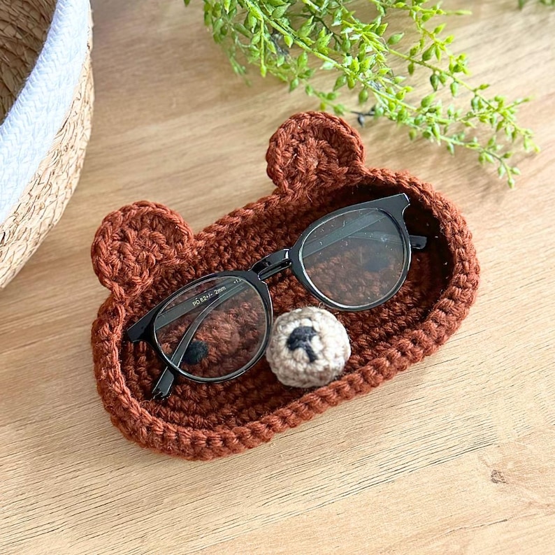May include: A brown crocheted bear-shaped glasses holder with a small crocheted nose. A pair of black framed glasses are resting inside the holder. The glasses have the text 'PD 6311-C 52□18-135' printed on the left arm.