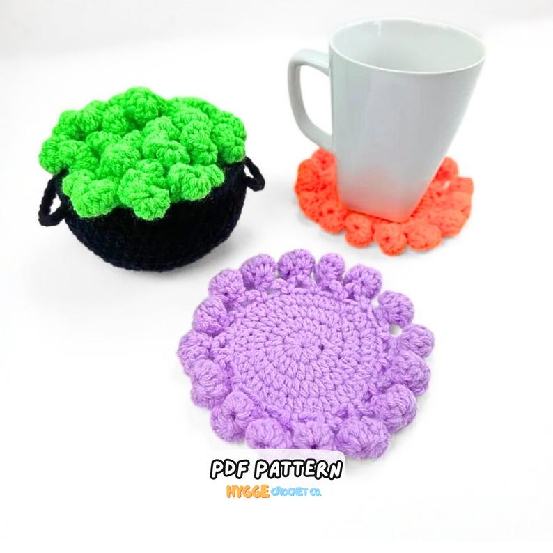 May include: A black crochet cauldron filled with bright green textured yarn, a white ceramic mug resting on an orange crochet coaster, and a separate purple crochet coaster with a scalloped edge. The text PDF PATTERN HYGGE CROCHET CO is visible.