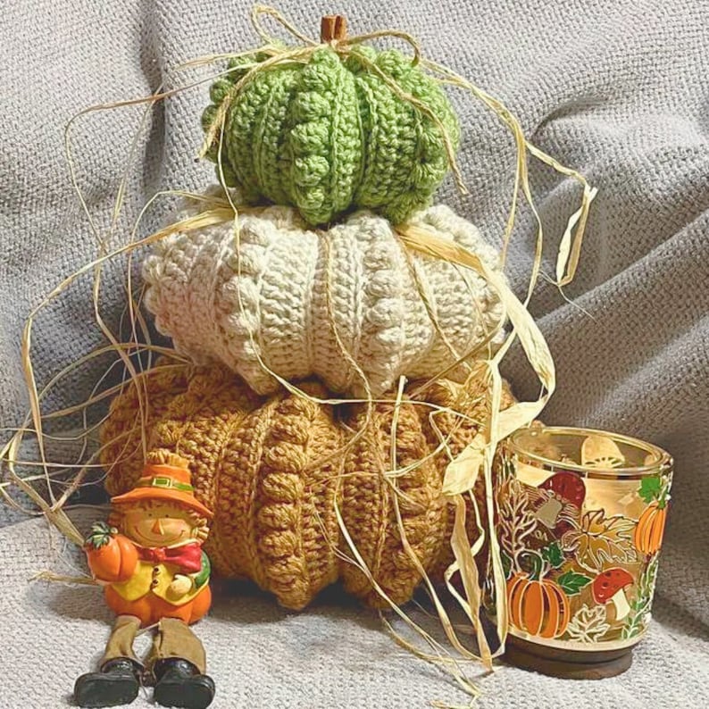 Puede incluir: Una pila de tres calabazas de ganchillo en verde claro, crema y amarillo mostaza, rematadas con una rama de canela y rafia. Junto a las calabazas hay una peque&ntilde;a figura de espantap&aacute;jaros y un portavelas de cristal con motivos oto&ntilde;ales.
