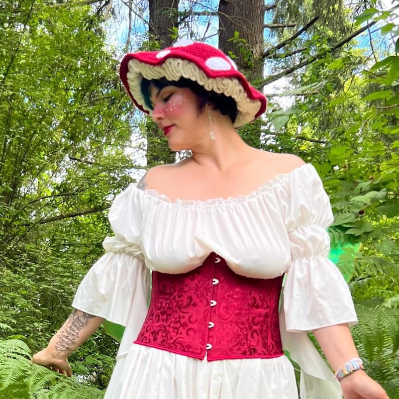 May include: A red and white crocheted mushroom hat with a ruffled cream-colored brim, paired with a white off-the-shoulder blouse featuring puffed sleeves and a deep red corset with a tonal damask pattern and a front row of silver-toned buttons.