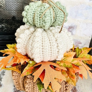 Puede incluir: Una decoraci&oacute;n de calabazas de ganchillo apiladas en tres niveles de color verde salvia, crema y bronceado. La parte superior est&aacute; acentuada con una rama de canela y un lazo de cuerda. La base est&aacute; rodeada de hojas de oto&ntilde;o artificiales.