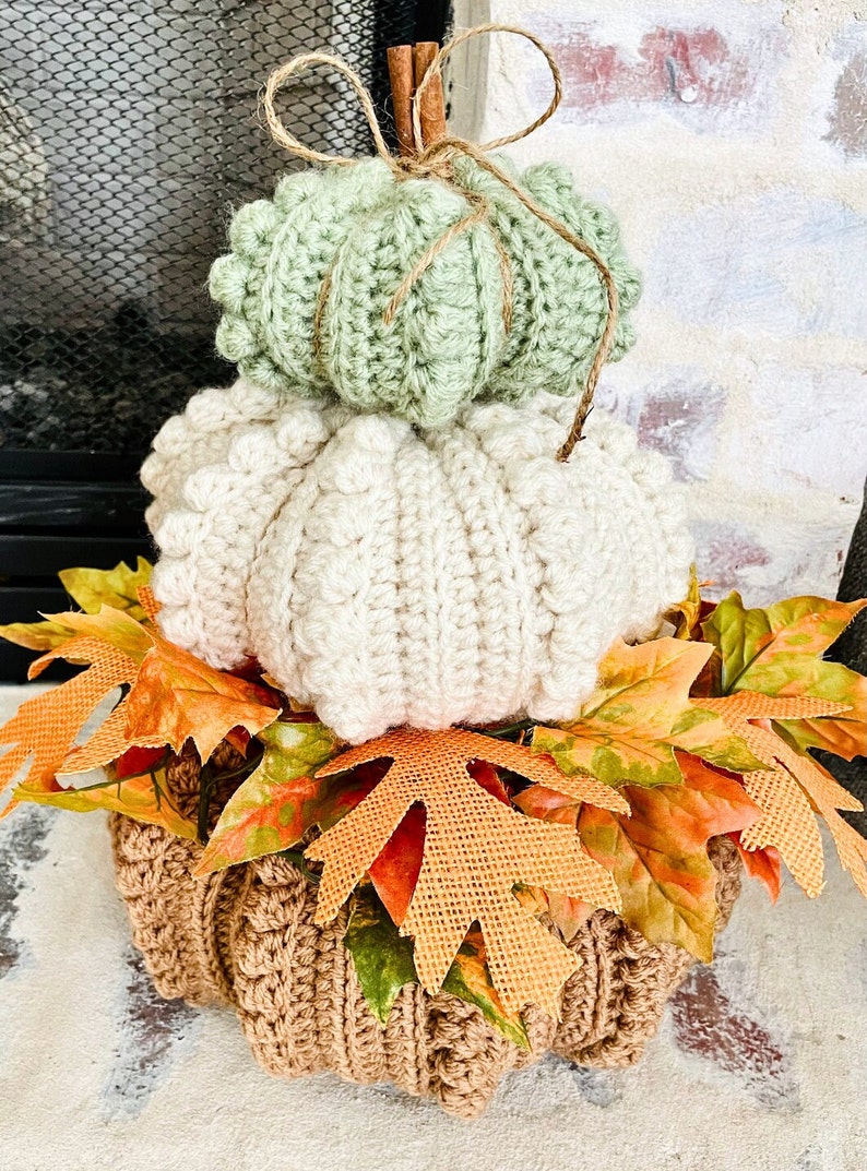Puede incluir: Una decoraci&oacute;n de calabazas de ganchillo apiladas en tres niveles de color verde salvia, crema y bronceado. La parte superior est&aacute; acentuada con una rama de canela y un lazo de cuerda. La base est&aacute; rodeada de hojas de oto&ntilde;o artificiales.