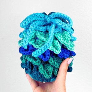 May include: A small, round, crocheted pouch featuring a textured, scale-like pattern in shades of blue, teal, and green. The top of the pouch is cinched closed with a matching light blue crocheted drawstring, creating a gathered, ruffled appearance.