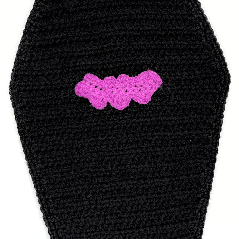 May include: A black coffin-shaped object crafted from crochet yarn, featuring a textured surface with horizontal stitching lines. Centered on the front is a small, bright pink crocheted bat applique with scalloped edges and a distinct wing shape.