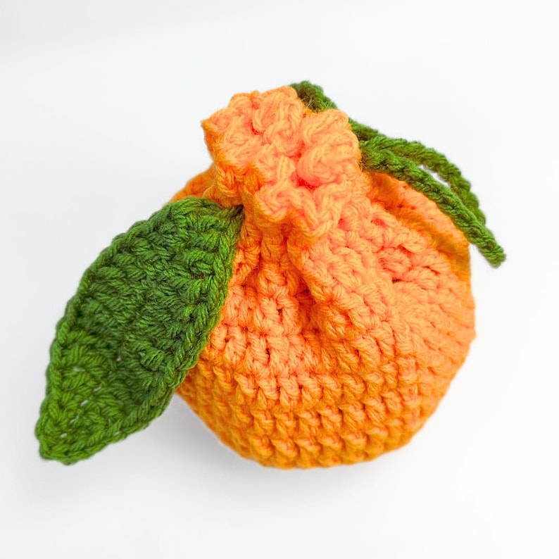 K&ouml;nnte beinhalten: Ein rundes, orangefarbenes H&auml;kelbeutelchen in Form einer Zitrusfrucht. Die Oberseite ist mit einem Kordelzug gerafft und mit zwei strukturierten, dunkelgr&uuml;nen H&auml;kelbl&auml;ttern an der Seite versehen, was ein realistisches Fruchtbild ergibt.