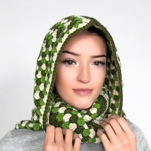 May include: A crochet hood and scarf combination featuring a granny square pattern in shades of dark green, light green, and cream. The accessory is constructed with a textured yarn stitch, forming a continuous piece that covers the head and neck.
