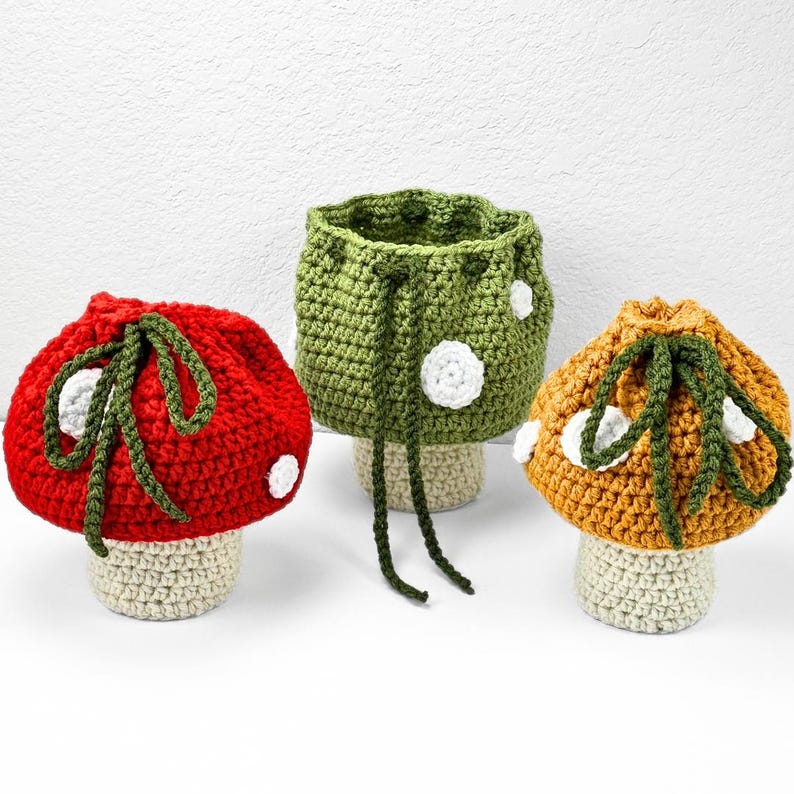 May include: Three crochet mushroom-shaped pouches with drawstring closures. The left pouch is red, the center is green, and the right is orange. Each features a beige stem base, dark green drawstring ties, and small white circular spots.