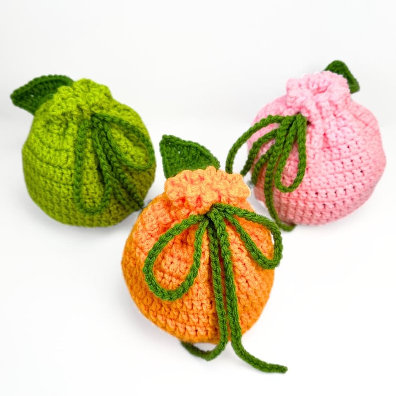 K&ouml;nnte beinhalten: Drei runde, geh&auml;kelte Beutel mit Kordelzug in Fruchtform. Einer ist limettengr&uuml;n, einer leuchtend orange und einer hellrosa. Jeder Beutel hat ein oben befestigtes, dunkelgr&uuml;nes geh&auml;keltes Blatt und einen dunkelgr&uuml;nen Kordelzug zum Verschlie&szlig;en.