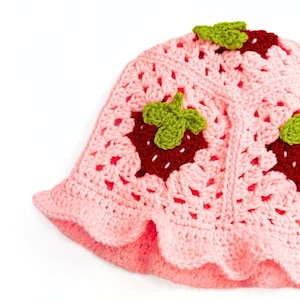 K&ouml;nnte beinhalten: Ein rosa geh&auml;kelter Hut mit Erdbeer-Applikationen. Der Hut hat eine breite Krempe und ist mit gr&uuml;nen Bl&auml;ttern und roten Beeren verziert.