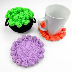 May include: A black crocheted cauldron filled with bright green yarn bubbles, a white ceramic mug resting on an orange crocheted coaster with a scalloped edge, and a separate round purple crocheted coaster featuring a textured, bobble-stitch border.