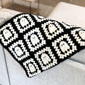 May include: A crochet blanket made of square motifs featuring a white ghost design with black eyes on a black background. The blanket is constructed from multiple repeating squares joined together, creating a high-contrast pattern of white ghosts.