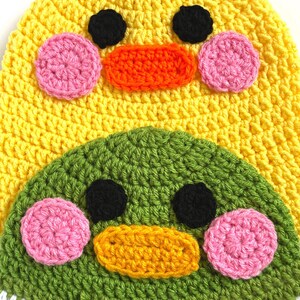 Pu&ograve; includere: Un cappello all'uncinetto con un design di anatra giallo e verde. L'anatra ha occhi neri, guance rosa e un becco arancione. Il cappello ha una striscia bianca e marrone nella parte inferiore.