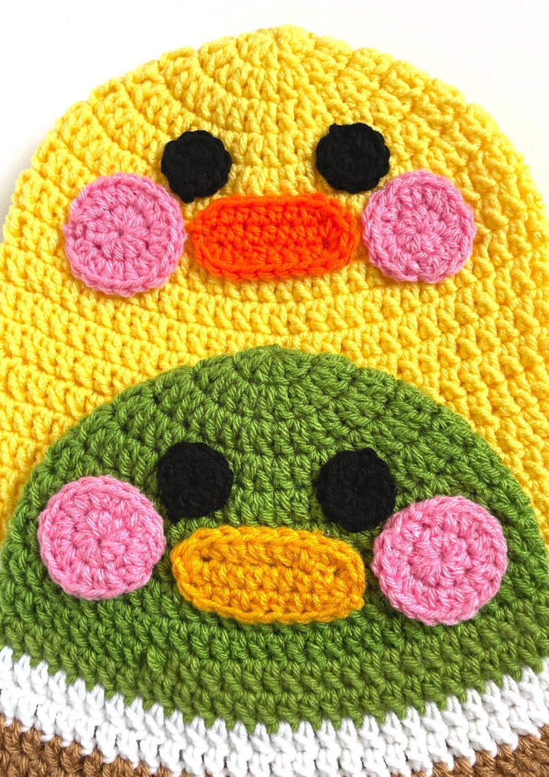 Pu&ograve; includere: Un cappello all'uncinetto con un design di anatra giallo e verde. L'anatra ha occhi neri, guance rosa e un becco arancione. Il cappello ha una striscia bianca e marrone nella parte inferiore.