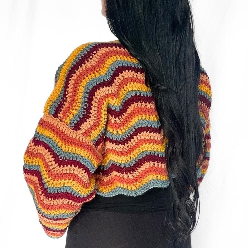 May include: A long-sleeve crochet sweater featuring a wavy, multi-colored striped pattern in shades of mustard yellow, deep burgundy, light orange, and muted blue. The garment has a cropped length and a textured, hand-stitched knit construction.