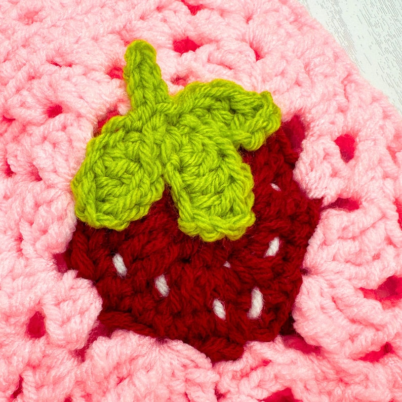 K&ouml;nnte beinhalten: Nahaufnahme einer geh&auml;kelten Erdbeere mit einem gr&uuml;nen geh&auml;kelten Blatt. Die Erdbeere ist rot und das Blatt ist gr&uuml;n. Die Erdbeere befindet sich auf einem rosa geh&auml;kelten Hintergrund.