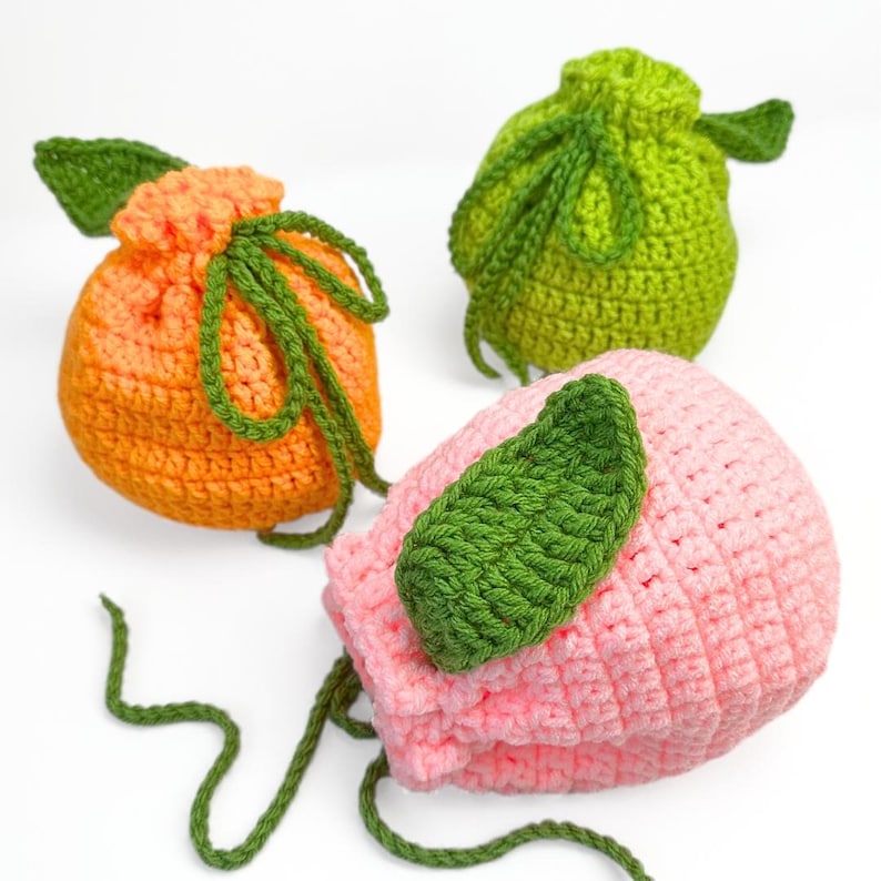 K&ouml;nnte beinhalten: Drei geh&auml;kelte Beutel mit Kordelzug in Fruchtform. Einer ist orange mit gr&uuml;nem Blatt und B&auml;ndern, einer ist limettengr&uuml;n mit gr&uuml;nem Blatt und B&auml;ndern, und einer ist hellrosa mit gr&uuml;nem Blatt und langen gr&uuml;nen Kordelzugb&auml;ndern.