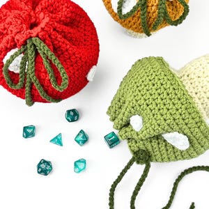May include: Three crocheted mushroom-shaped drawstring pouches in red, orange, and green, each featuring white spots and green stems. Scattered among the pouches are six translucent teal polyhedral dice with white numbering.