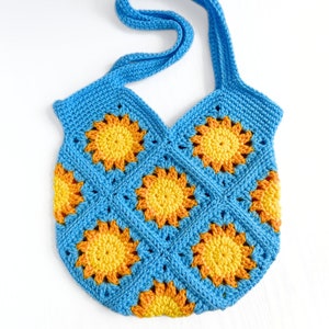 May include: A crochet tote bag constructed from square motifs featuring a bright blue border and a central sunburst pattern in shades of yellow and orange. The bag includes a single integrated shoulder strap and a textured, hand-stitched finish.