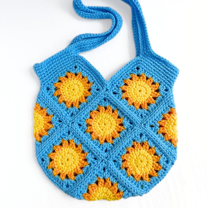 May include: A crochet tote bag constructed from square motifs featuring a bright blue border and a central sunburst pattern in shades of yellow and orange. The bag includes a single integrated shoulder strap and a textured, hand-stitched finish.