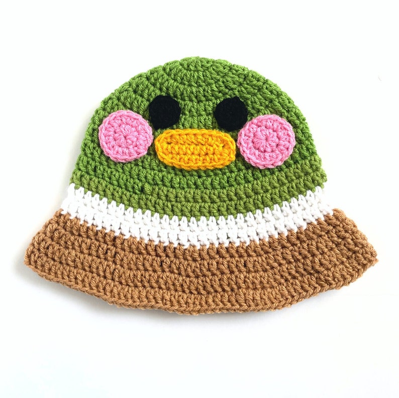 Pu&ograve; includere: Cappello a secchiello in crochet verde con un viso di papera da cartone animato. La papera ha le guance rosa, gli occhi neri e un becco giallo.
