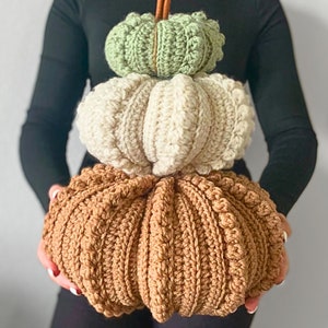 Puede incluir: Una pila de tres calabazas tejidas a ganchillo en orden descendente de tama&ntilde;o, con una calabaza verde salvia arriba, una crema en el medio y una grande de color marr&oacute;n abajo. Cada calabaza tiene una superficie texturizada y acanalada.