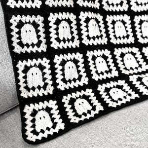 May include: A handmade crochet blanket featuring a repeating pattern of square motifs. Each square consists of a white ghost figure centered within a black and white border. The textile is constructed from interconnected yarn squares.