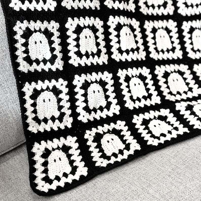 May include: A handmade crochet blanket featuring a repeating pattern of square motifs. Each square consists of a white ghost figure centered within a black and white border. The textile is constructed from interconnected yarn squares.