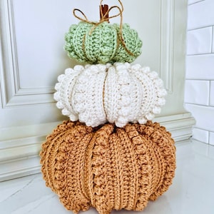 Puede incluir: Una pila de tres calabazas tejidas a ganchillo en orden descendente de tama&ntilde;o. La calabaza inferior es naranja, la del medio es crema y la superior es verde claro. Cada calabaza presenta un patr&oacute;n de puntos texturizados y un tallo de canela.