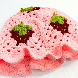 K&ouml;nnte beinhalten: Rosa gestrickte M&uuml;tze mit einem gewellten Rand und drei gestickten Erdbeeren. Die Erdbeeren sind rot mit gr&uuml;nen Bl&auml;ttern.