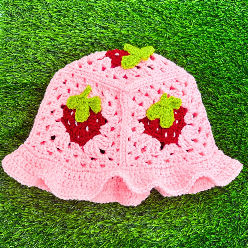 K&ouml;nnte beinhalten: Ein rosa geh&auml;kelter Sonnenhut mit Erdbeer-Design. Der Hut hat eine breite Krempe und einen gewellten Rand.