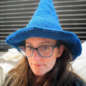 May include: A blue crocheted witch hat with a wide brim. The hat is made with a textured stitch and has a pointed crown.