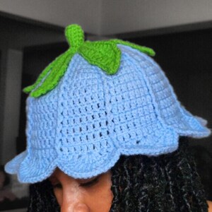 May include: A light blue crochet hat shaped like a bell flower with a scalloped edge. The top of the hat features a bright green crocheted stem and leaves. The hat is constructed with a textured stitch pattern that mimics the petals of a flower.