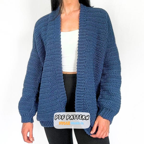 May include: A dark blue open-front crochet cardigan with long sleeves and ribbed cuffs. The garment features a textured stitch pattern throughout. A white label with black text reads PDF PATTERN and orange text reads HYGGE.