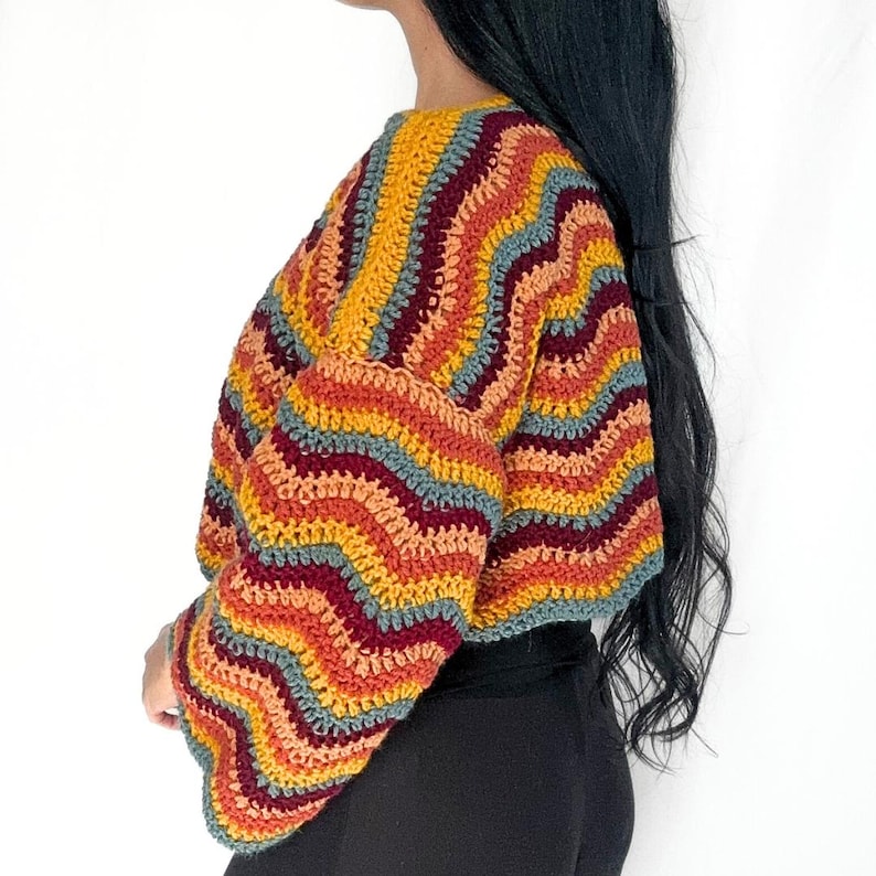 May include: A long-sleeved crochet sweater featuring a wavy, multi-colored striped pattern in shades of mustard yellow, burnt orange, deep burgundy, and muted teal. The garment has a round neckline and a cropped fit, showcasing a textured knit.