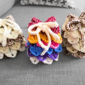 May include: Three small drawstring pouches crafted with a textured crocodile stitch pattern. The center pouch features a vibrant rainbow gradient of red, orange, yellow, blue, and purple yarn, while the two side pouches are in neutral brown and cream tones.