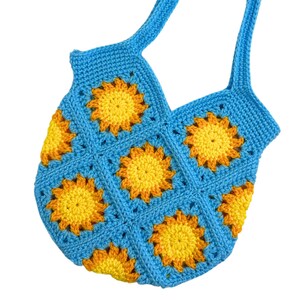 May include: A handmade crochet tote bag constructed from multiple square motifs. Each square features a bright yellow and orange sunburst pattern centered within a light blue border. The bag includes two integrated shoulder straps in blue.